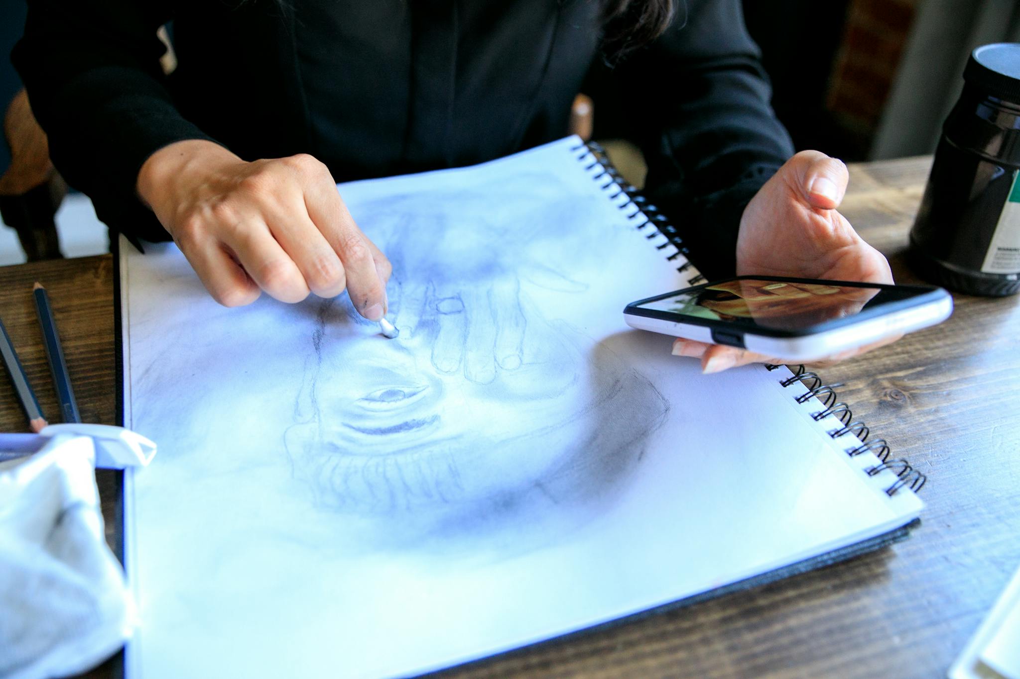 An artist uses a smartphone while adding details to a pencil drawing in a sketchbook.